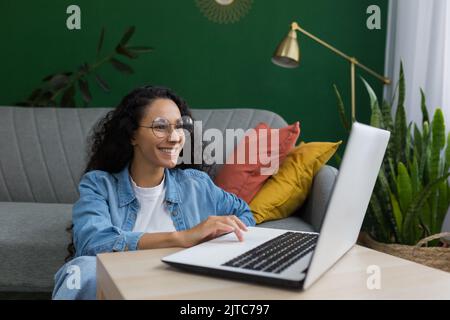 Young beautiful female student studying remotely online using laptop, Latin American woman sitting on floor at home in green living room, watching online course lecture Stock Photo
