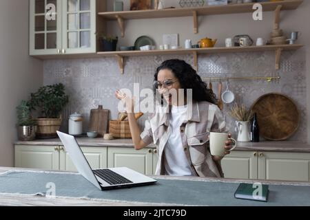 Young beautiful hispanic housewife celebrating victory and joyfully looking at laptop screen, woman with curly hair working remotely in kitchen at home Stock Photo