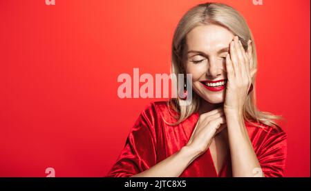 Beautiful middle aged woman with blond hair, stylish makeup, lauging and smiling, touching her flawless skin, standing over red background Stock Photo