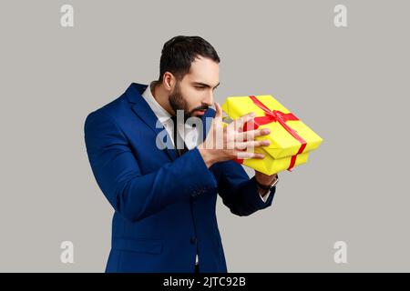 Handsome bearded man unpacking present, looking inside box with curious serious expression, birthday gist, wearing official style suit. Indoor studio shot isolated on gray background. Stock Photo