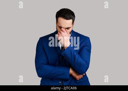 Sad upset unhappy bearded businessman hiding face in hand crying, feeling stressed worried, expressing sorrow, wearing official style suit. Indoor studio shot isolated on gray background. Stock Photo