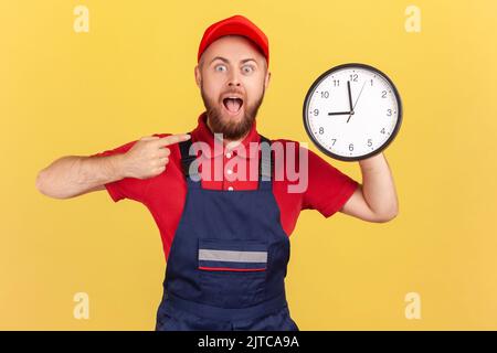 Shocked surprised man worker standing and pointing at big wall clock in his hands, looking at camera with open mouth, wearing overalls and red cap. Indoor studio shot isolated on yellow background. Stock Photo