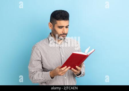 Concentrated bearded businessman holding book and reading, standing absorbed with exciting plot, checking schedule in notepad, wearing striped shirt. Indoor studio shot isolated on blue background. Stock Photo