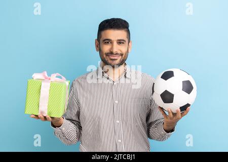 Portrait of happy delighted businessman standing looking smiling at camera, holding soccer ball and present box, wearing striped shirt. Indoor studio shot isolated on blue background. Stock Photo