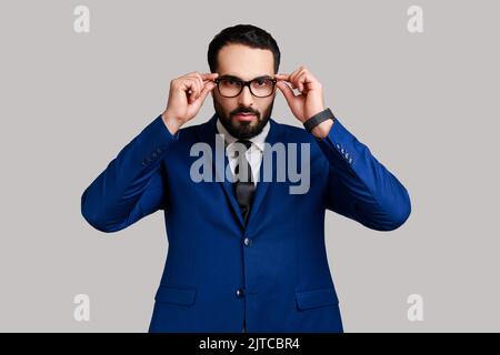 Portrait of serious bearded businessman putting off eyeglasses and looking at camera with bossy expression, wearing official style suit. Indoor studio shot isolated on gray background. Stock Photo