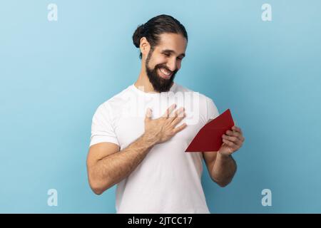 Portrait of man with beard wearing T-shirt reading letter from red envelope, gets greeting card, having positive romantic expression, being pleased, Indoor studio shot isolated on blue background. Stock Photo