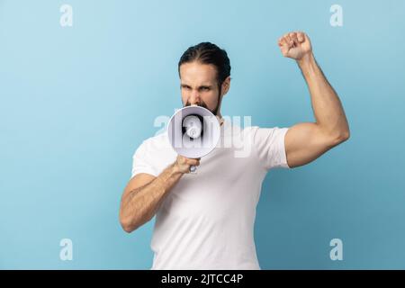 Portrait of angry man with beard wearing white T-shirt loudly screaming at megaphone, making announce, protesting, wants to be heard. Indoor studio shot isolated on blue background. Stock Photo