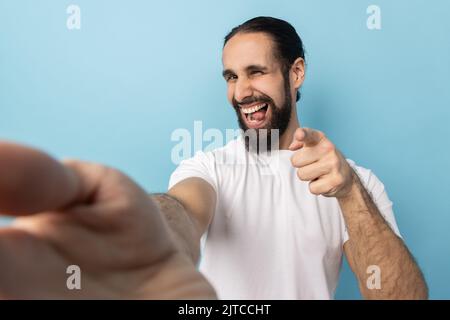 Portrait of satisfied man with beard wearing white T-shirt looking at camera with tongue out and pointing finger on you, taking selfie POV. Indoor studio shot isolated on blue background. Stock Photo