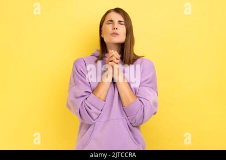 Portrait of young attractive woman keeps eyes closed imploring desperate grimace, praying to god asking for help, wearing purple hoodie. Indoor studio shot isolated on yellow background. Stock Photo