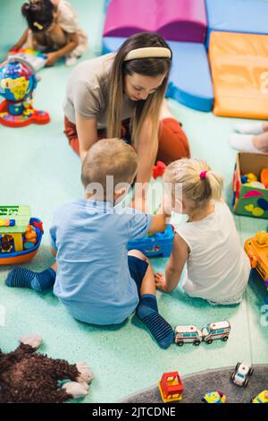 Learning through play at the nursery school. Caucasian toddlers and their teacher playing with colorful plastic playhouses, building blocks, cars and boats. Imagination, creativity, fine motor and gross motor skills development. High quality photo Stock Photo