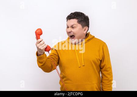 Portrait of nervous man screaming and yelling talking retro landline phone, complaining on connection quality, wearing urban style hoodie. Indoor studio shot isolated on white background. Stock Photo