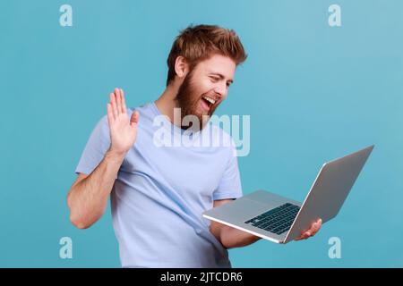 Portrait of friendly bearded man greeting a worker through a webcam and showing palm of the hand gesture with toothy smile, having online conversation. Indoor studio shot isolated on blue background. Stock Photo