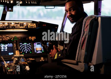 Portrait of african american pilot using dashboard command in cabin to takeoff with plane jet and fly. Male aviator in airways uniform pushing control panel buttons and power lever. Stock Photo