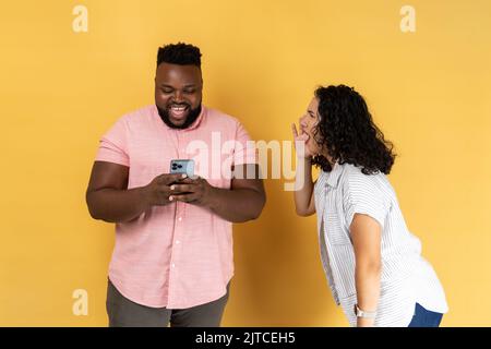 Young couple in casual clothing standing together, woman desperately screaming near happy man using phone and ignoring her, attracting his attention. Indoor studio shot isolated on yellow background. Stock Photo