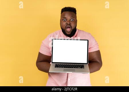 Portrait of shocked amazed man wearing pink shirt holding notebook, showing white blank screen on his laptop, looking at camera with big eyes. Indoor studio shot isolated on yellow background.