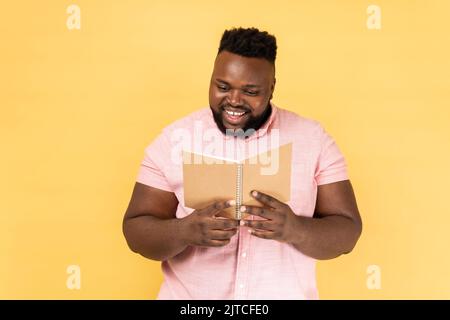 Portrait of delighted pleasant looking man wearing pink shirt holding book and reading interested novel, expressing positive emotions. Indoor studio shot isolated on yellow background. Stock Photo