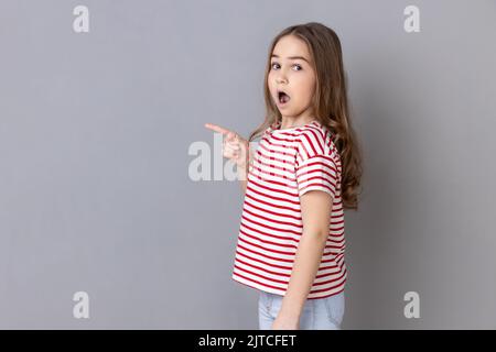 Wow, look at crazy advertisement. Side view of little girl wearing striped T-shirt pointing aside at empty place for ad content and expressing surprise. Indoor studio shot isolated on gray background Stock Photo