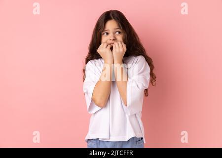 Troubles and worries. Portrait of little girl wearing white T-shirt biting nails, terrified about problems, suffering phobia, anxiety disorder. Indoor studio shot isolated on pink background. Stock Photo