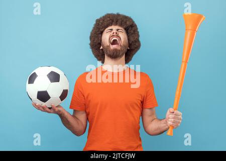 Man with Afro hairstyle wearing T-shirt screaming widely opening mouth, celebrating victory of favourite football team, holding horn and ball in hands. Indoor studio shot isolated on blue background. Stock Photo