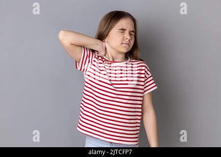 Portrait of unhealthy sick dark haired little girl wearing striped T-shirt standing massaging neck to relieve pain, muscle strain in back. Indoor studio shot isolated on gray background. Stock Photo