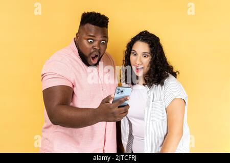 Amazed young couple in casual clothing standing together and reading unexpected message with open mouth, using mobile phone, checking social networks. Indoor studio shot isolated on yellow background. Stock Photo