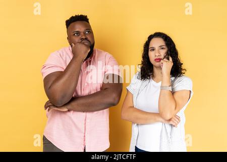 Portrait of thoughtful young couple in casual clothing standing together, holding their chins while thinking solution, looking away. Indoor studio shot isolated on yellow background. Stock Photo