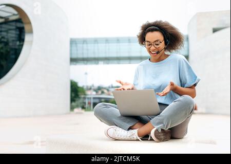 Positive friendly african american curly young woman with headset, manager, coach, having online consultation, negotiations with colleagues, sits outdoors, using laptop, talking on video call, smiling Stock Photo