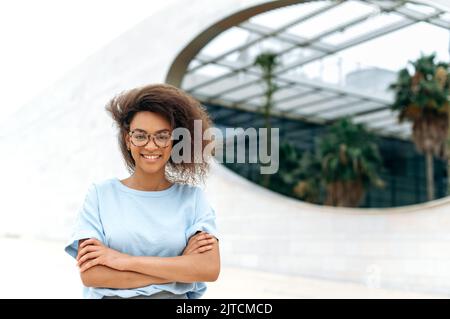 Photo of successful pretty African American curly young business woman, in elegant stylish clothes, with glasses, stands outdoors near business center with arms crossed, looks at camera, smiles Stock Photo