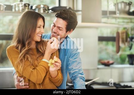 Want some. an attractive young woman feeding her boyfriend grapes in their kitchen. Stock Photo