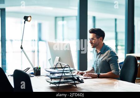 True success comes from doing. a focused young businessman using a computer at his desk in a modern office. Stock Photo
