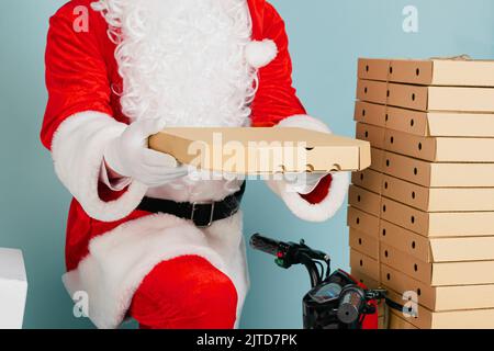 A closed pizza box close-up in the hands of a santa claus holds in his hands against the background of a red moped with a stack of pizza boxes on a bl Stock Photo