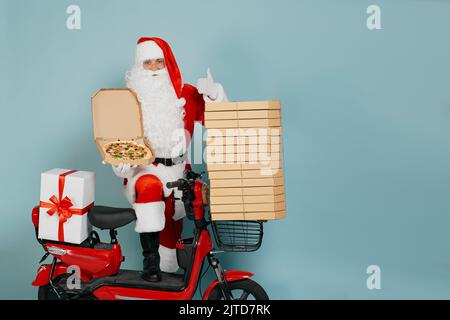 Santa claus demonstrates pizza in an open box close-up on the background of a red moped with a stack of boxes on a blue isolated background with copy Stock Photo