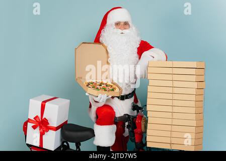 Pizza delivery man dressed as santa claus with an open pizza box in his hand cloaks himself on a stack of boxes on the background of a red scooter and Stock Photo