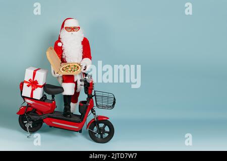 Full length portrait of santa claus in big red heart shaped glasses with an open box of pizza held in his hands in white gloves, against a red moped o Stock Photo