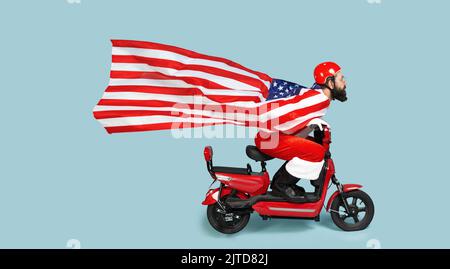 Santa with a big flying usa flag in the form of a raincoat and a helmet rides a red electric scooter on a blue isolated background with copy space Stock Photo