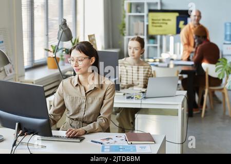 Portrait of young woman using computer at desk in office and smiling with people working in background Stock Photo