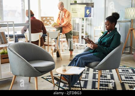 Side view portrait of modern black businesswoman using digital tablet in office with people working in background, copy space Stock Photo