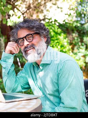 Bearded middle aged man smiling happy on terrace working from home using laptop Stock Photo