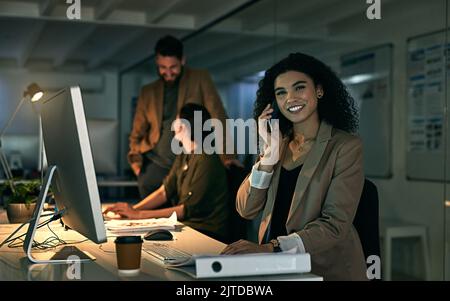 She has no problem putting in extra time for clients. a young woman talking on her phone with her colleagues in the background. Stock Photo