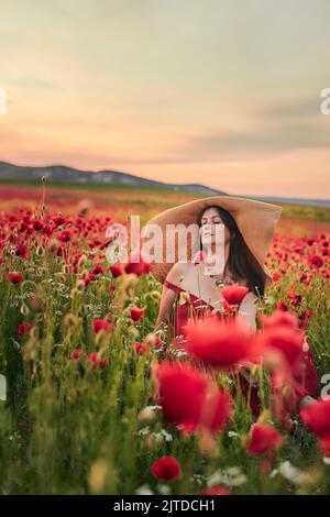 Portrait of a woman in a poppy field wearing a wide-brimmed hat at sunset. Vertical photo. Stock Photo