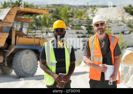 Two multicultural male workers of marble quarry standing in front of camera against dump truck on territory of industrial factory Stock Photo