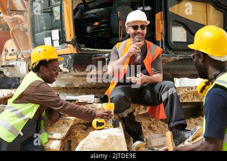 Group of happy middle aged intercultural workers of marble quarry having lunch and communicating while sitting by excavator Stock Photo