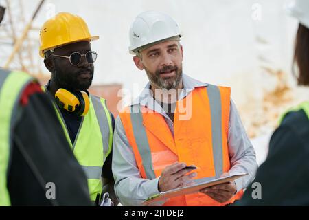Confident foreman with document and pen explaining something to subordinates or giving instructions while looking at one of them Stock Photo