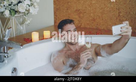 Handsome young man is taking selfie with champagne glass using smartphone in hot tub with foam in modern spa salon. He is smiling and posing looking at camera. Stock Photo