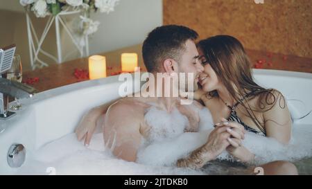 Young attractive lovers in bathing suits are hugging and kissing in jacuzzi with foam in day spa. Romantic relationship, passion, relaxation and wellness concept. Stock Photo