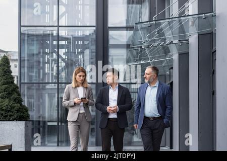 Diverse business group, three male and female workers walking and chatting discussing plans, outside office building Stock Photo
