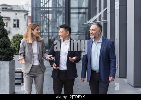 Diverse business group, three male and female workers walking and chatting discussing plans, outside office building Stock Photo