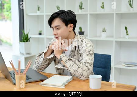 Thoughtful young Asian man office worker in casual outfit planning and working on his project on notebook laptop in his office. Stock Photo