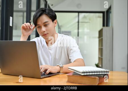 Smart young Asian male college student concentrating doing his work project homework in campus library, researching some information on internet and t Stock Photo