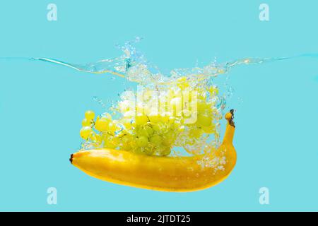 Bunch of white grapes and yellow banana dropped in water with splashes isolated on blue background. Stock Photo